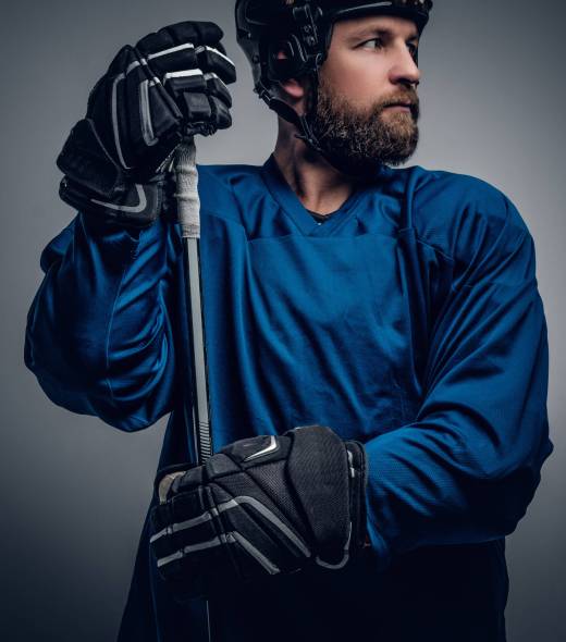 A brutal bearded ice-hockey player in safety helmet holds the gaming stick on grey vignette background.